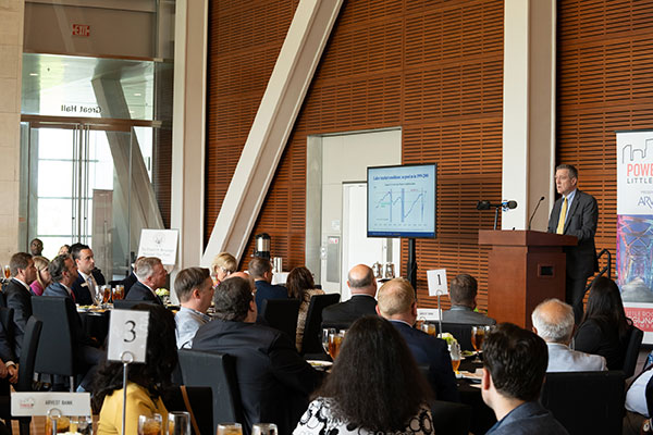 Man in business attire stands at a lectern in front of a seated crowd in business attire.