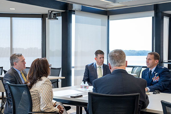 Four men and a woman in business attire sit at a conference table overlooking water. 