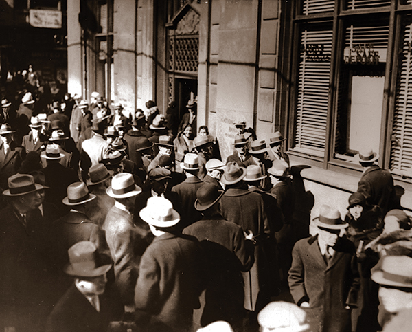 Black and white photo of people in hats, lined up outside of a bank.