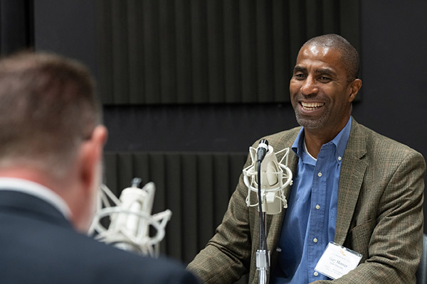 A seated man wearing a sportcoat smiling in front a microphone.