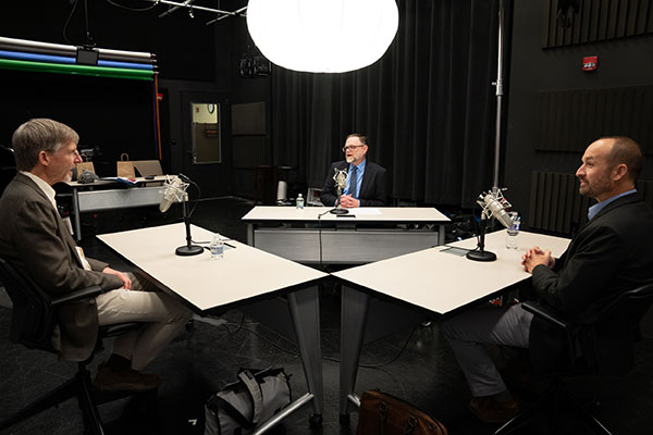 Three men in business attire sit in a podcast recording studio.