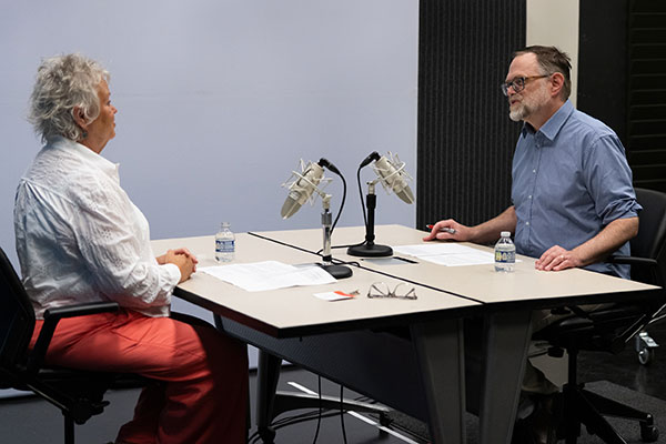 Two people sitting across from each other at a table set up for recording with individual microphones.