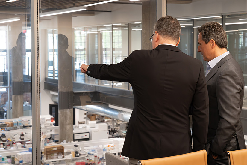 A man stands with Musalem and gestures at a room with shelves of bottles on the other side of a glass wall.