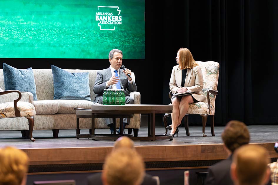 Musalem speaks to an audience while seated on a couch next to a woman in a chair. A green screen behind them has ‘Arkansas Bankers Association’ text on an Arkansas map.