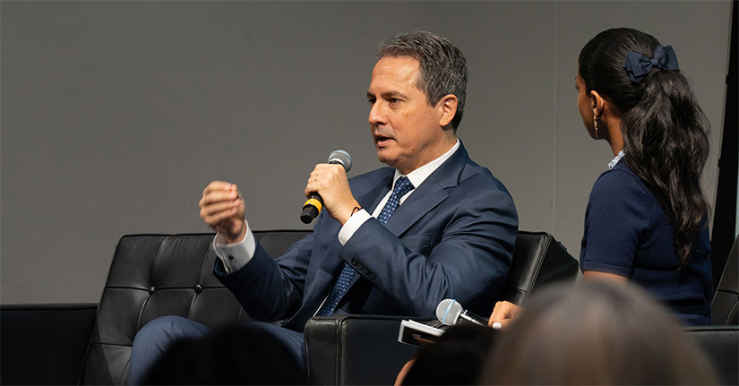 Musalem speaks into a microphone while seated next to a woman on a stage in front of audience members.