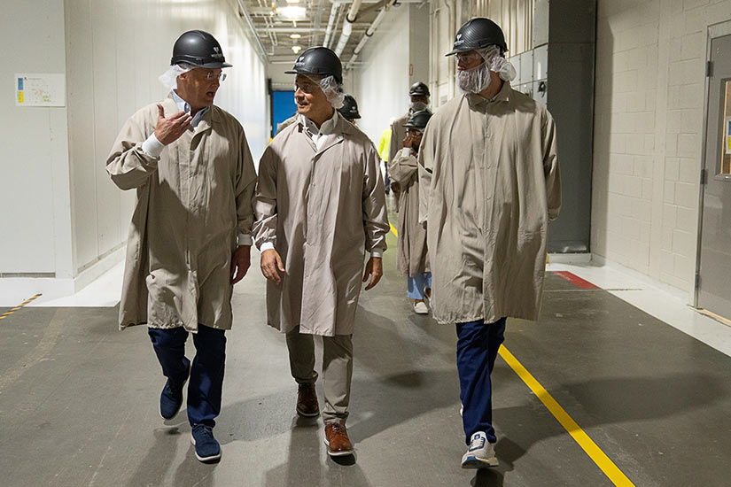 Musalem walks down a large hallway with two men and others in the background. All wear protective smocks, hair covers and helmets.