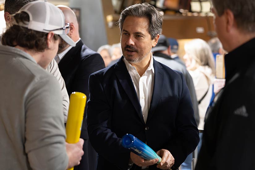 A man in business attire holding a blue baseball bat talks with a man in a ballcap holding a yellow baseball bat.