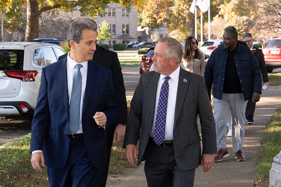 St. Louis Fed President Alberto Musalem and a group of people in business attire walk on a sidewalk along a tree-lined street.