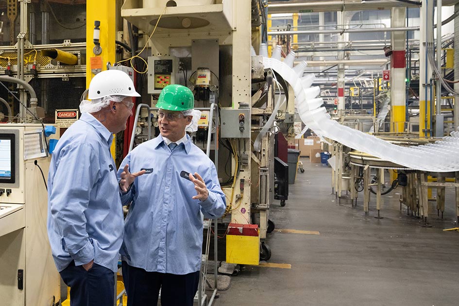 President Musalem gestures while speaking to another man as they stand in a factory in front of a machine rolling out plastic. Both men are wearing hard hats and blue collared shirts.