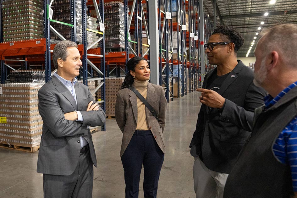 President Musalem and a woman and a man in business attire listen to a man as they stand in a warehouse in front of high metal shelves filled with packages.