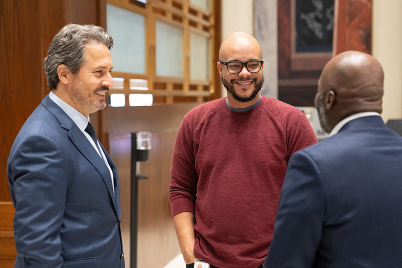 Three men smile in conversation in a large hallway.