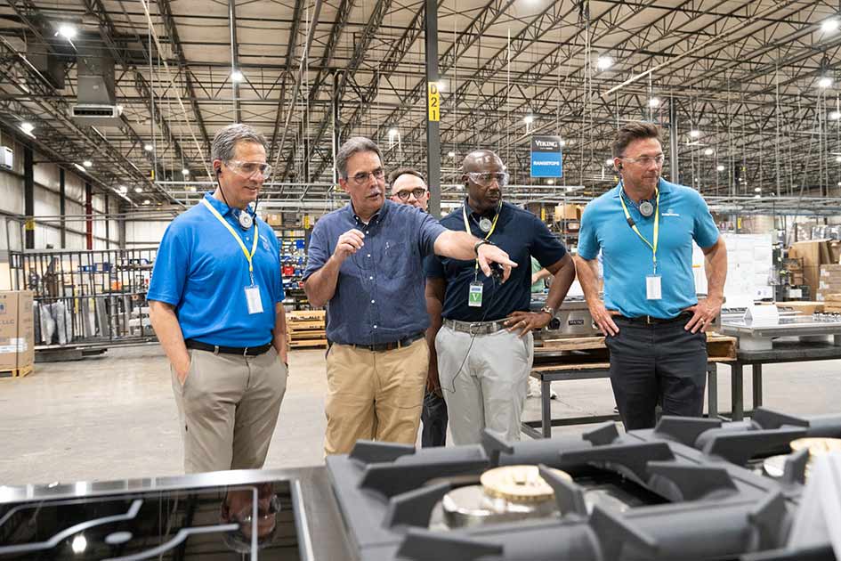 Standing in a manufacturing facility, St. Louis Fed President Alberto Musalem and three others watch as a man gestures toward kitchen appliances.