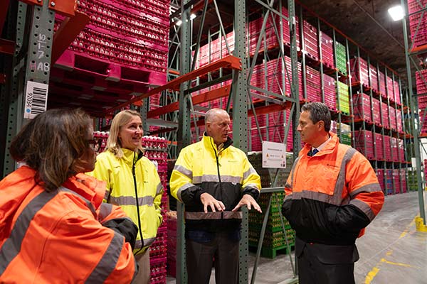 A man gestures while speaking to President Musalem and two women, all in bright yellow or orange jackets, in front of high metal shelves filled with cartons of eggs in a warehouse.