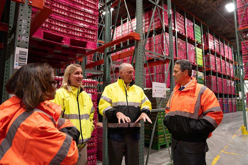 A man gestures while speaking to President Musalem and two women, all in bright yellow or orange jackets, in front of high metal shelves filled with cartons of eggs in a warehouse.