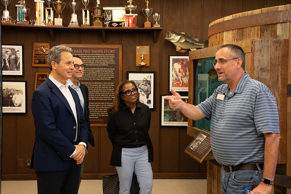 President Musalem and another man and a woman in business attire listen to a man in casual attire while standing in a room with plaques and photos on the wall.