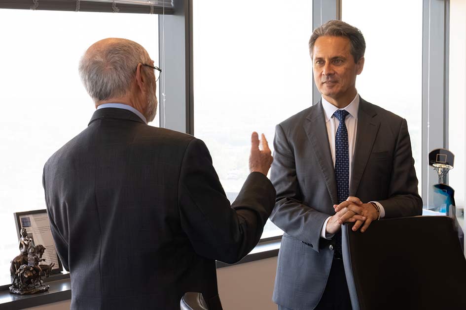 A man with his back to the camera speaks to St. Louis Fed President Alberto Musalem in front of office windows.