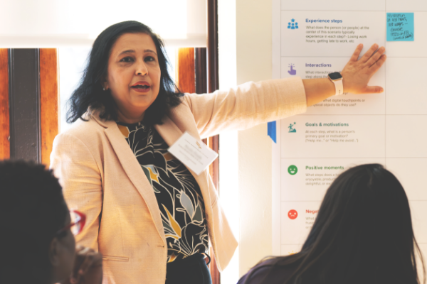 A woman points to a whiteboard as she speaks to a group.
