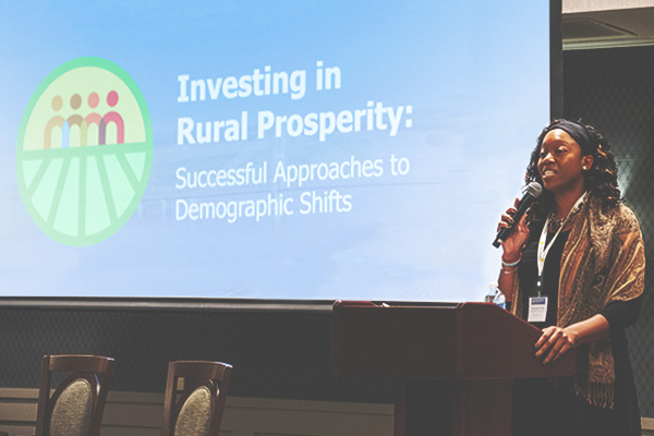 Speaker presents to an audience in front of screen displaying 'Investing in Rural Prosperity: Successful Approaches to Demographic Shifts.'