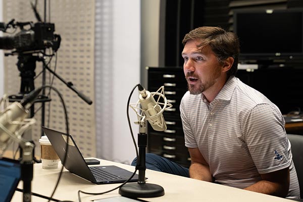 A man in business attire speaks in front of a microphone in a studio.