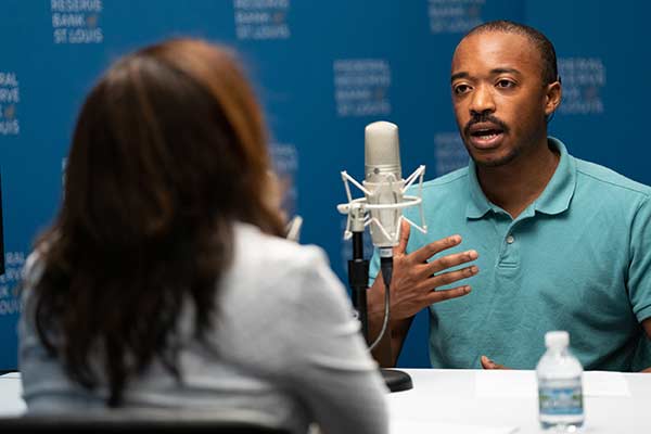 A man and women in business attire have a discussion in front of microphones in a podcast studio.