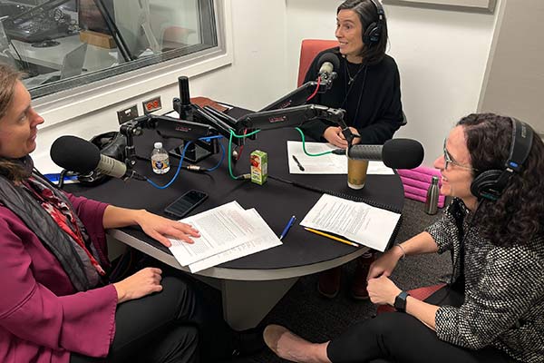 Three women sit at a table with headphones and microphones in discussion.
