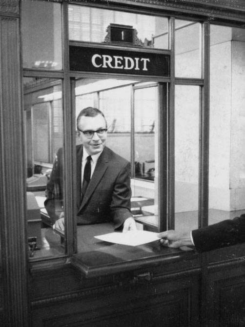 Photo of a man in a business suit handing a sheet of paper to unseen person from a bank teller window labeled credit.