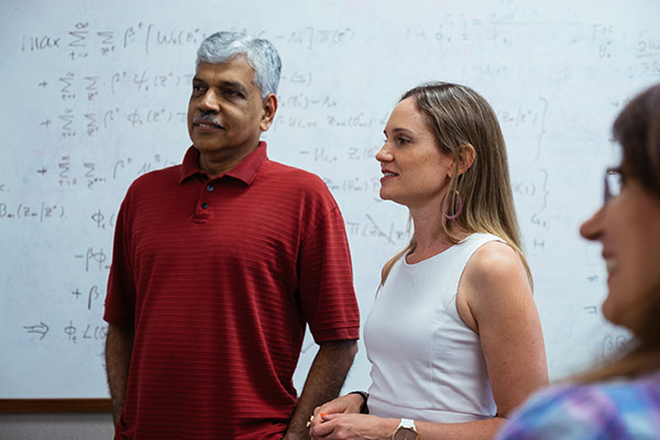 Three economists engage in conversation in front of a white board.