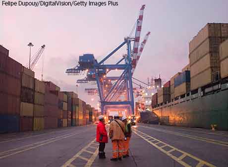 A group of workers talking among themselves on a dock, where a ship is being loaded with cargo.